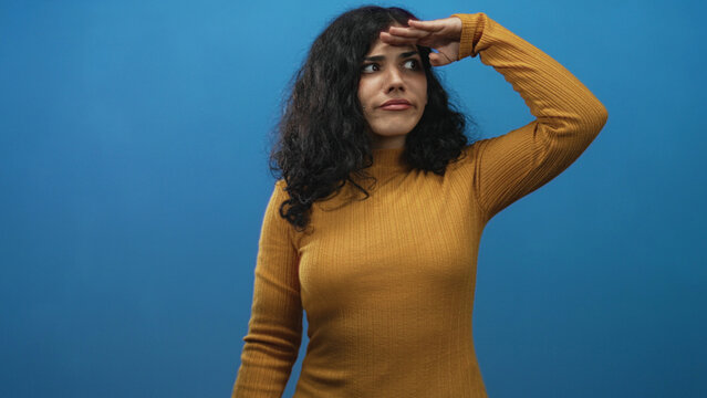Young arab woman shielding eyes with hand while scanning the distance in a blue studio wearing a mustard sweater and curly hair; curiosity searching.