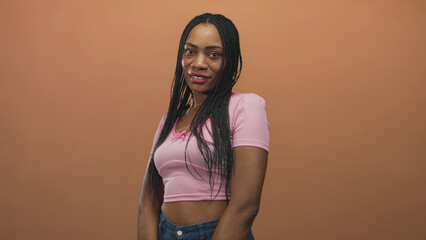 Obraz premium Woman with braided hair showing midriff, smiling and slightly tilting her head in a studio wearing a pink top and jeans; confidence warmth.