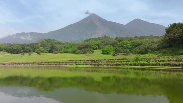 Active Fuego volcano emits smoke above lush tropical forest in Guatemala. Calm lake reflects vibrant green hills and majestic volcanic peaks under hazy blue sky in Central America.