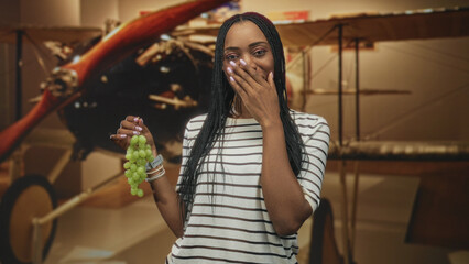 Woman holding a bunch of green grapes with hand covering mouth beside a vintage biplane inside museum building  surprise curiosity. © Krakenimages.com