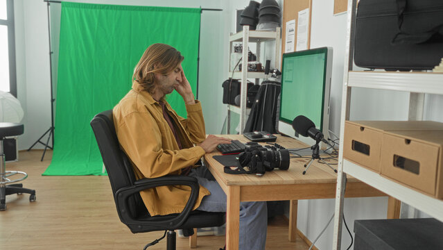 Man photographer types on keyboard with hand to face at computer in studio; concentration creativity.