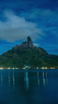 Vertical Screen: Mount Otemanu rises majestically against deep blue twilight sky in Bora Bora. Coastal lights shimmer along shoreline with reflections dancing on calm lagoon waters beneath stars.