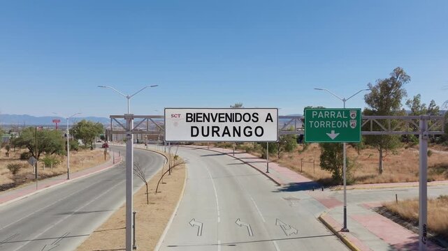 Aerial drone footage of Welcome to Durango sign on highway in Mexico with direction signs to Parral and Torreon. Clear blue sky over arid landscape with scattered trees and empty roads.