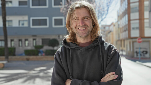 Man with long blond hair and arms crossed, smiling with hands visible, wearing a dark hoodie while standing on a sunny urban street in front of buildings; everyday contentment.