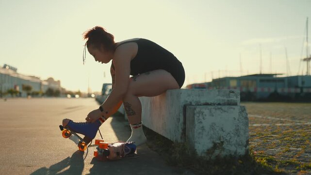 Young tattooed woman tying roller skates outdoors at sunset