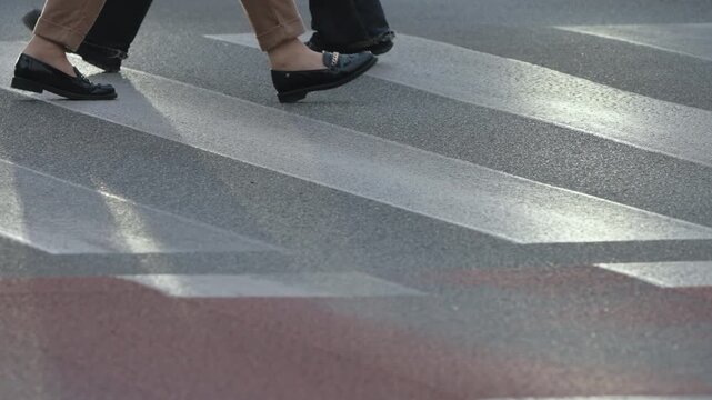 Cinematic close up view urban crosswalk with zebra stripes on asphalt, pedestrians walking in slow motion and bicycles passing intermittently, legs to knee level, overcast cloudy city atmosphere