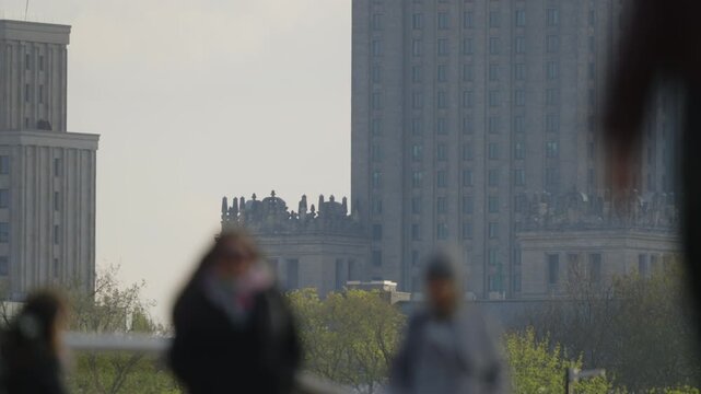 Urban lifestyle scene telephoto compression and shallow depth of field, pedestrians and cyclists crossing sidewalk in foreground blur, sunlight, distant high-rise building facade in background