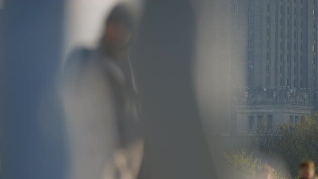 City scene with shallow depth of field, cyclists and pedestrians moving across sidewalk foreground blur, strong telephoto compression revealing distant skyline and tall landmark facade under daylight