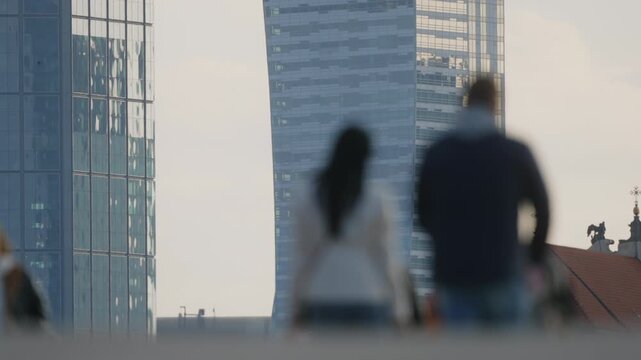 Urban lifestyle scene telephoto compression and shallow depth of field, pedestrians and cyclists crossing sidewalk in foreground blur, sunlight, distant high-rise building facade in background