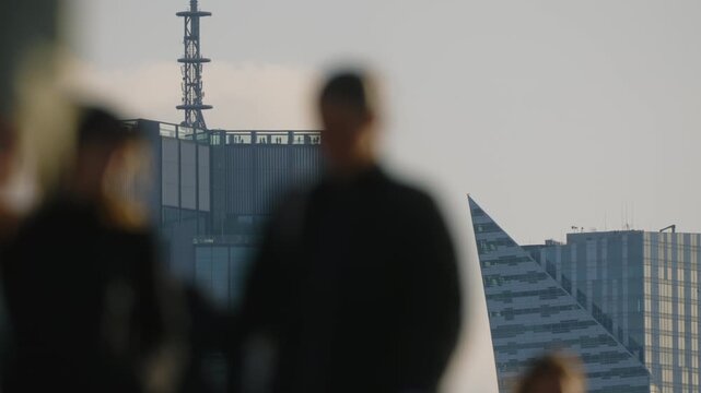 Urban skyline glass skyscrapers under soft morning or evening light, observation deck with people visible on rooftop in distance, blurred pedestrians moving in foreground, calm city lifestyle scene