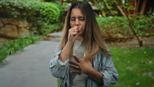 Hispanic woman coughing into fist clutching chest for heartattack in green park pathway; health concern distress.