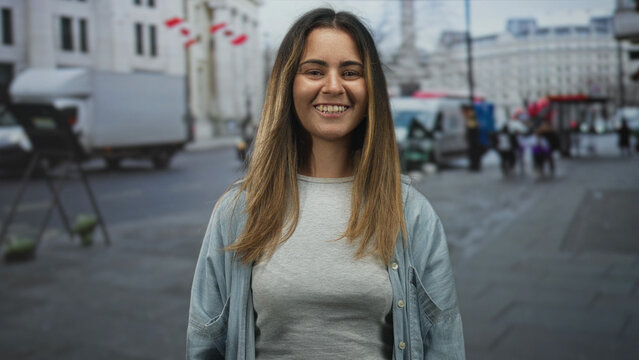 Woman smiling with eyes closed showing face and teeth in casual denim shirt on busy city street with vans and pedestrians; happiness calm optimism.