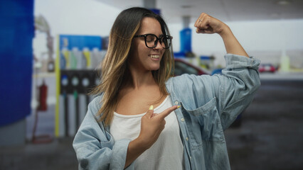 Hispanic woman flexes bicep and points to arm at petrol station forecourt wearing glasses and denim shirt  empowerment confidence. © Krakenimages.com