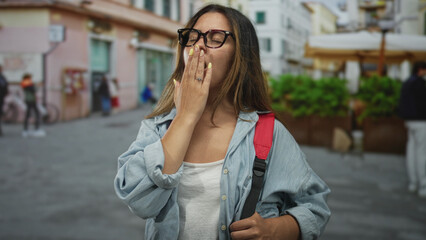 Obraz premium Woman covering mouth with hand and eyes closed while yawning on a city street with backpack visible; sleepiness.