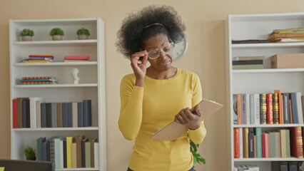 Woman in yellow sweater holding a clipboard and flipping pages while smiling and adjusting glasses beside packed bookshelves in building  contentment focus. © Krakenimages.com