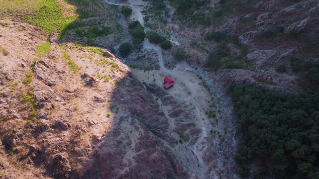 Aerial drone footage captures solitary red-roofed building nestled in a rugged mountain canyon with winding dirt roads, rocky terrain bathed in golden afternoon sunlight, and lush green forest.