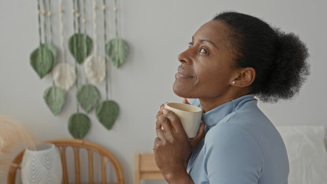 Woman holds coffee cup to chest with closed eyes on bed by rattan headboard and leaf wall hanging in studio; morning serenity.