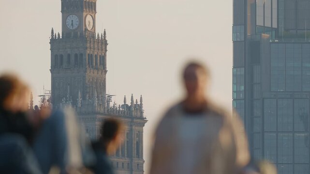 Urban lifestyle scene defocused foreground silhouettes on pedestrian bridge, people cycling and walking, distant skyline of glass skyscrapers and clock tower in focus, warm evening light, spring city