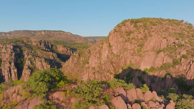 Aerial drone footage captures stunning red rock canyon formations bathed in warm golden hour sunlight, with rugged cliffs dotted by green vegetation under a clear blue sky in Sierra Madre mountains.