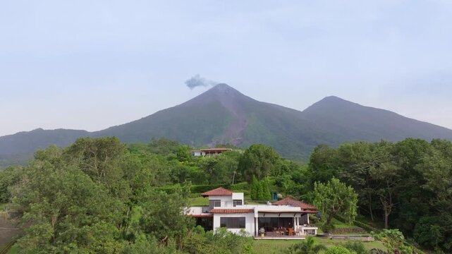 Aerial drone footage of a modern white villa with terracotta roof nestled in lush green tropical forest with active Fuego volcano emitting smoke in the misty background under overcast skies.