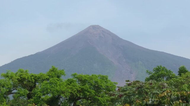 Guatemala active Fuego Volcano releases smoke plume from its peak while lush green tropical trees frame the majestic cone-shaped mountain against a hazy blue sky in Central America.