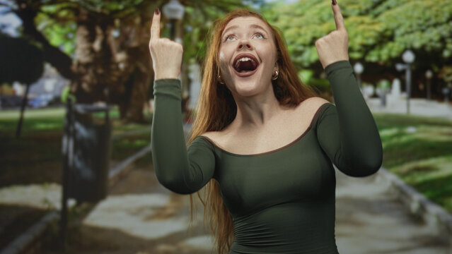 Woman pointing both index fingers upward with open mouth, visible hands and earrings on a street lined with trees and a trash bin; joy celebration.