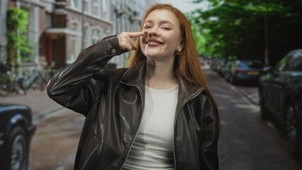 Fototapeta premium Woman pointing finger to nose on a street in front of parked cars and buildings, wearing leather jacket and smiling broadly; confidence openness.
