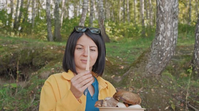 Caucasian woman camping testing knife blade tip for sharpness holds blade upright, calm focused look, preparing to clean wild mushroom collection, camp chef role, birch trees and soft daylight