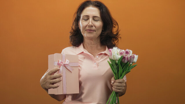 Middle age hispanic woman holds pink gift box and tulip bouquet, tilts head back with closed eyes in studio; quiet gratitude.