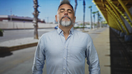 Fototapeta premium Man with gray beard and open collar standing with eyes closed, breathing slowly on a sunny street lined with palm trees and yellow canopy; calm reflection.