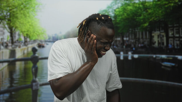 Man with hand on cheek for toothache on street canal bridge wearing white shirt and teeth clenched; pain struggle.