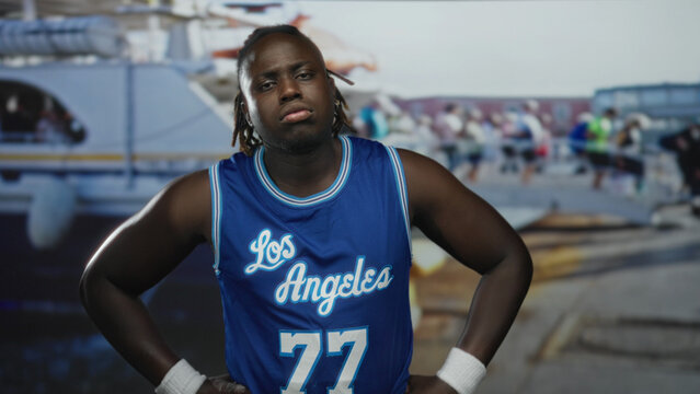 Man with hands on hips and bare forearms in blue basketball jersey on street pier near boats and dock; contemplation.