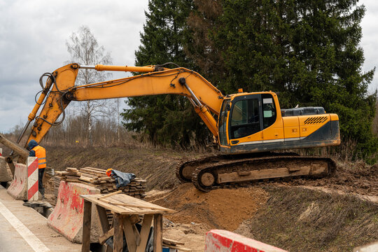 Yellow Excavator Suspended Over Embankment With Raised Bucket, Hydraulic Arm Extended, Tracked Undercarriage On Slope, Evergreen Backdrop, Precision