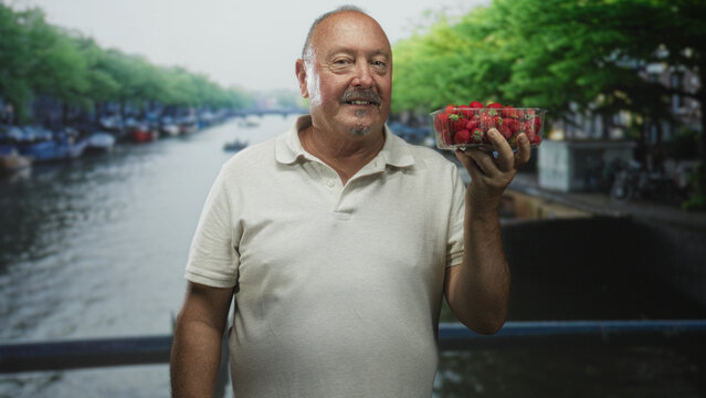 Man holding a punnet of strawberries on a street beside a canal bridge, smiling with relaxed posture; summer market contentment.