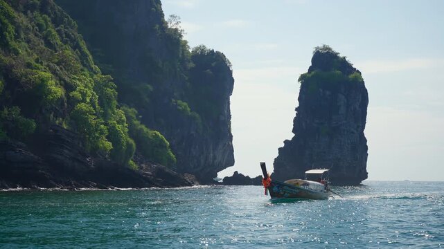 Traditional Thai longtail boat with colorful ribbons glides across shimmering turquoise waters beneath towering limestone karst cliffs covered in lush green vegetation under a hazy afternoon sky.