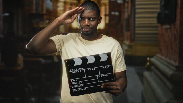 Man salutes with right hand while holding a clapperboard in a narrow urban street with shuttered storefronts and stone steps; confidence cinema.