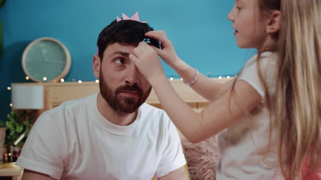 closeup pretty girl in dress putting hairpins and crown on his father's head making him a lovely male princess. father and daughter dressing for a costume party at home.