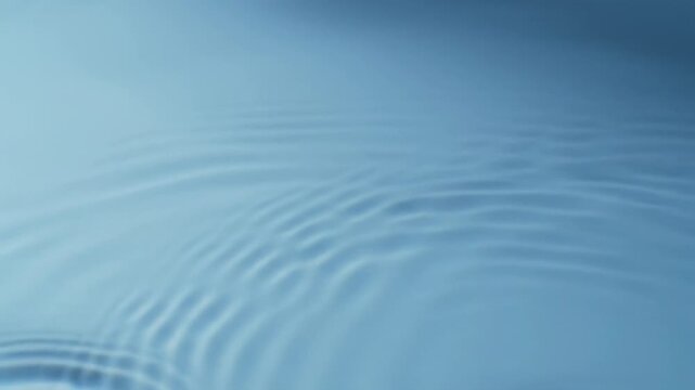 Pure overhead wide video of a pristine, crystal-clear water surface with subtle organic ripples and iridescent caustic light patterns on a diffused light blue background, executing an extremely slow