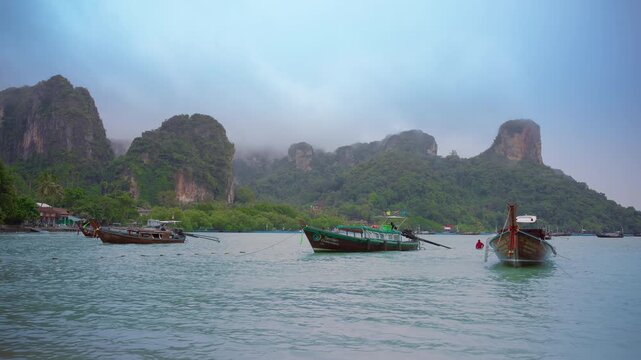 Traditional longtail boats float in turquoise waters of Mueang Krabi, Thailand with dramatic limestone karst mountains rising through morning mist in the background. Tropical coastal scenery.