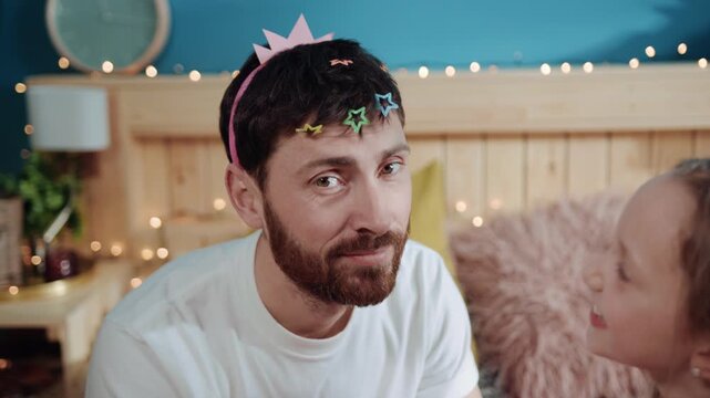 Portrait of miserable upset daddy crying for help while playing with his daughter. Poor tired father with hairpins in hair dressed like princess sitting with his child on bed.