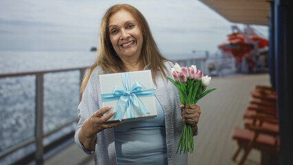 Fototapeta premium Woman holding a ribboned gift and a tulip bouquet on a cruise deck smiling gently; celebration joy gratitude.