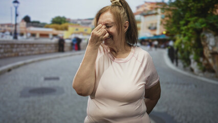 Fototapeta premium Woman pinches nose with fingers while standing on a cobblestone street beside a low stone wall and lamp post, eyes closed in a grimace; discomfort.