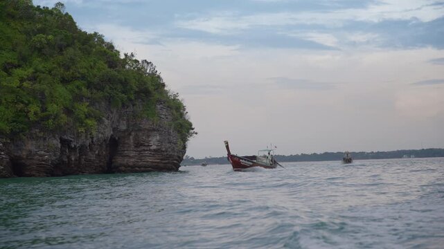 Traditional Thai longtail boats float on calm turquoise waters near a towering limestone cliff covered in lush green vegetation under an overcast evening sky in Krabi, Thailand, as gentle waves.