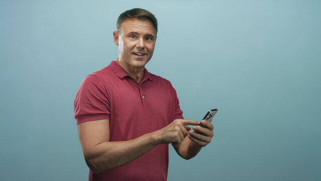 Man tapping smartphone with index finger and focused gaze in studio wearing red polo shirt, standing three quarter view; concentration connection.