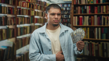 Man holding a fan of us dollars, hand on stomach for stomachache, gaze worried amid bookshelves in a library building  financial concern. © Krakenimages.com