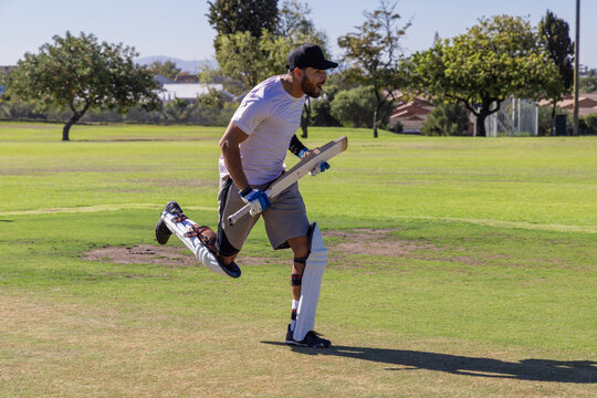 Wooden cricket bat is being carried while pads and gloves are glinting on grassy pitch