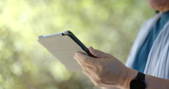 Senior Asian man holding folio tablet tapping screen in garden adjusting striped shirt, copy space