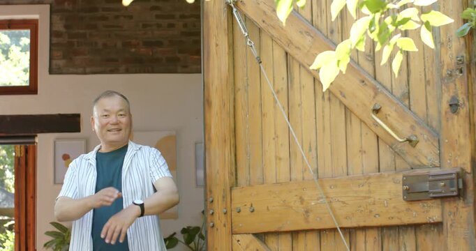 Senior Asian man hearing neighbor calling, stepping into home doorway, crossing arms by wooden gate