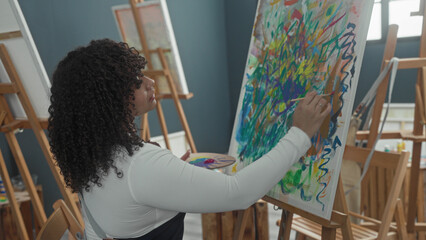 Woman painter with curly brunette hair seated at easel, painting colorful abstract canvas, holds...