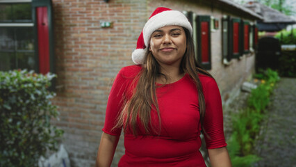 Fototapeta premium Woman wearing santa hat smiling and raises fists while swaying in red top in front of building with brick wall and window shutters; holiday joy.